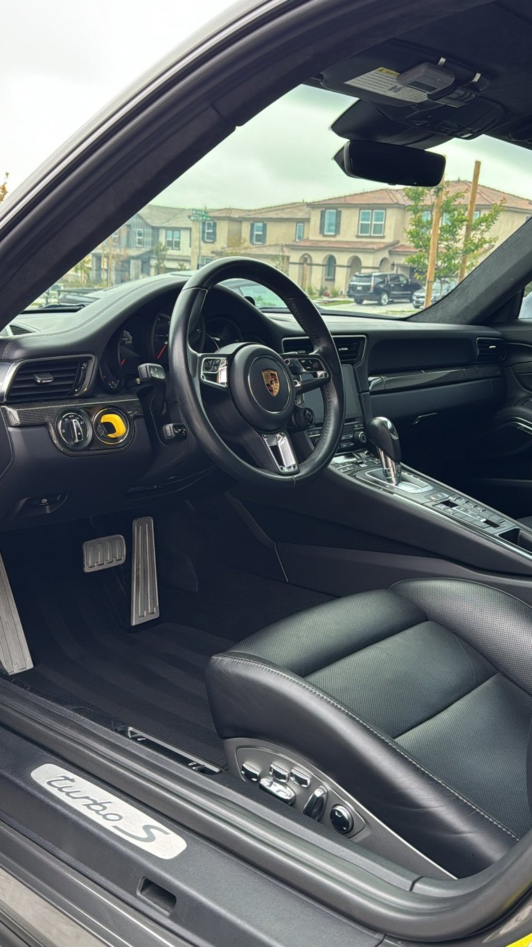 Interior view of a Porsche sports car with black steering wheel, dashboard, and leather seats, viewed through the open driver-side door